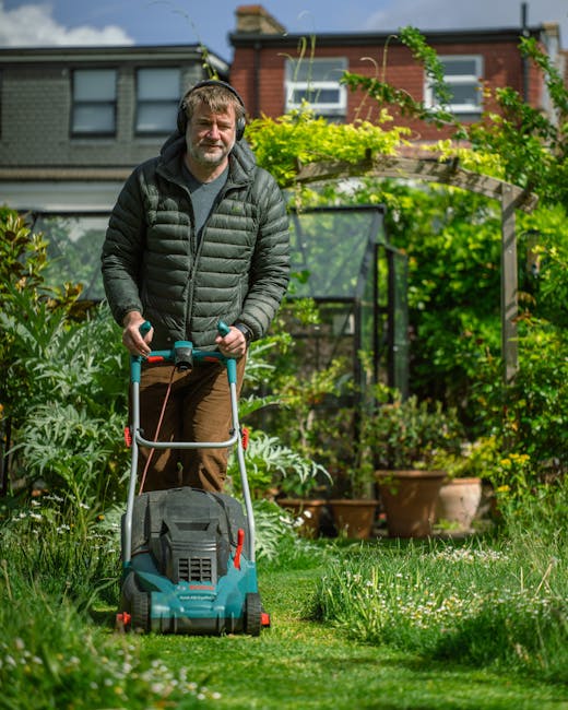 A man outdoors in a well-maintained garden, operating a petrol-powered lawn mower on a neatly trimmed grassy lawn. He has short, light-colored hair and a beard, and is wearing a dark puffer jacket and